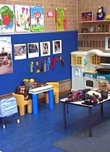 a room at a child care centre. The room has posters on the walls, and a variety of toys. The furniture is brightly coloured and child sized.